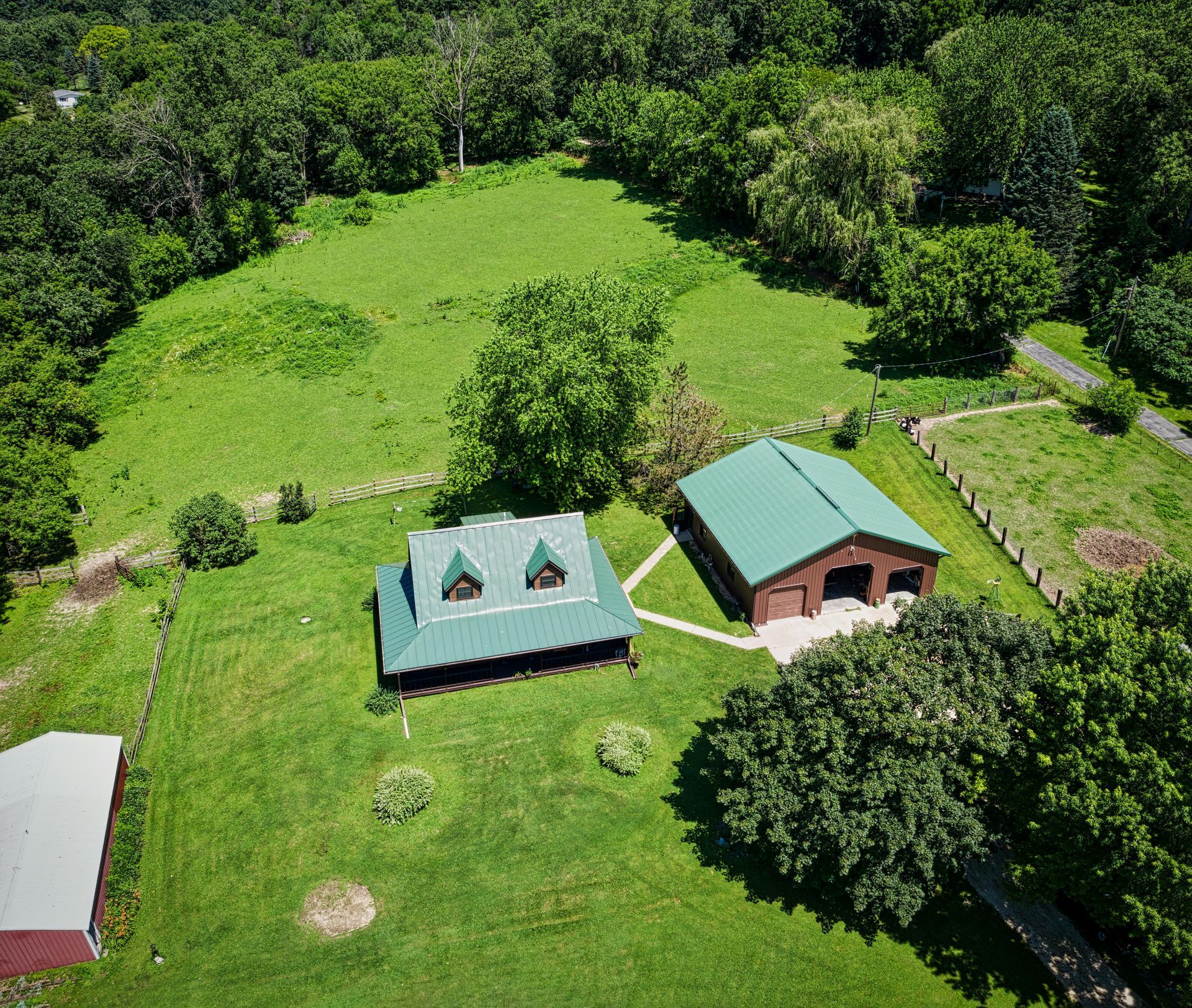 Aerial view of a green-roofed house and barn on a grassy property with trees and a fenced field.