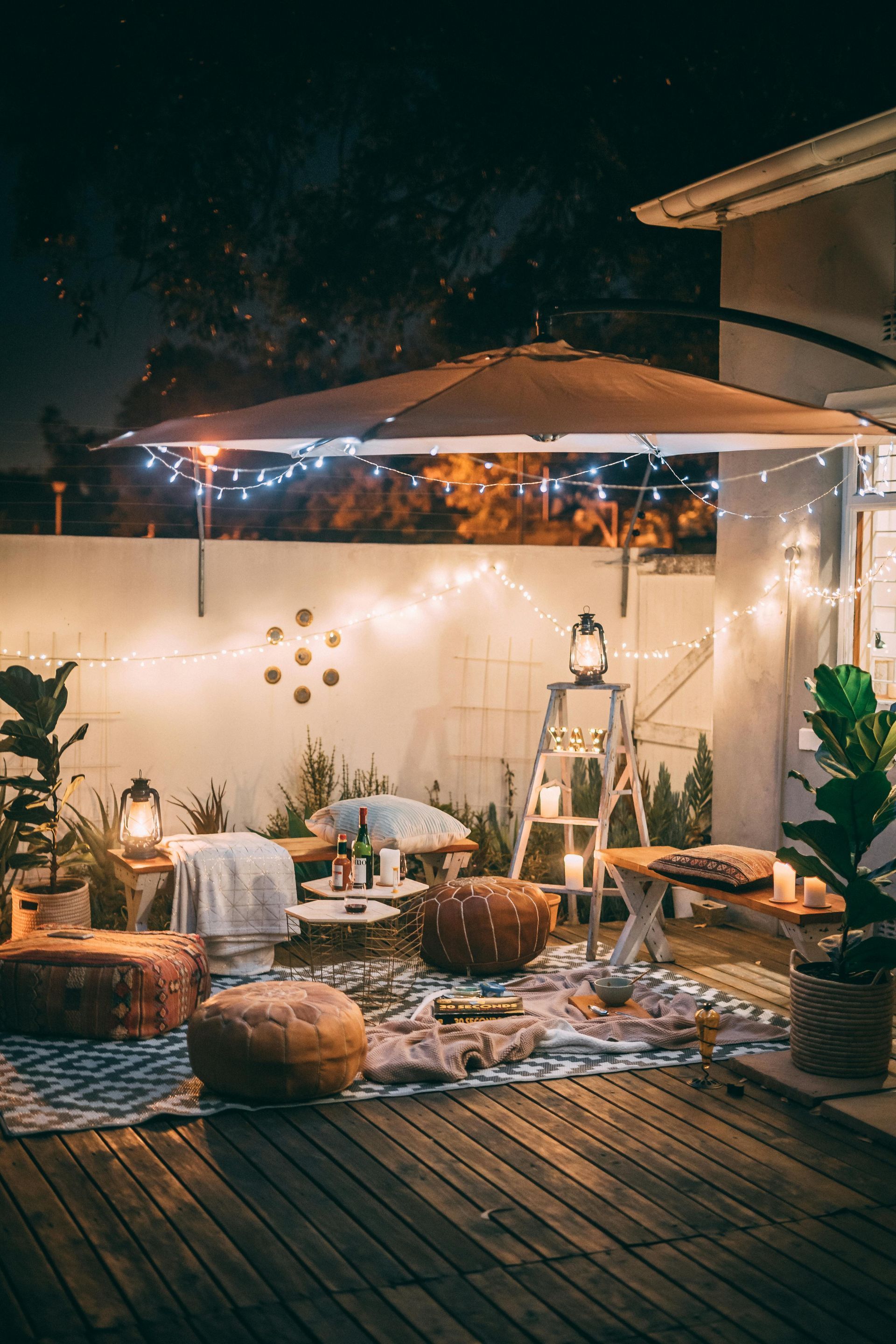 Cozy outdoor seating area with string lights, rugs, pillows, and a large umbrella at night.