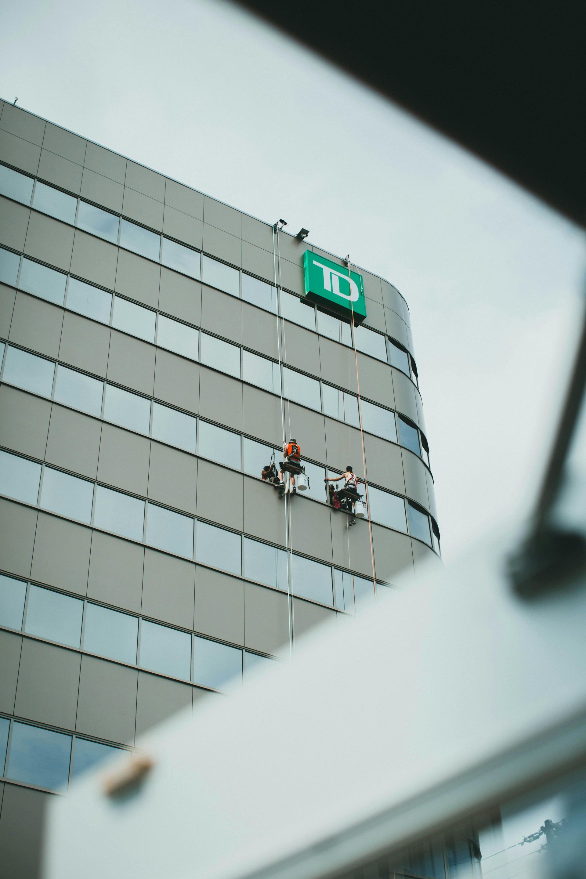 Two window washers on a tall TD Bank building, suspended by ropes. Cloudy sky above.