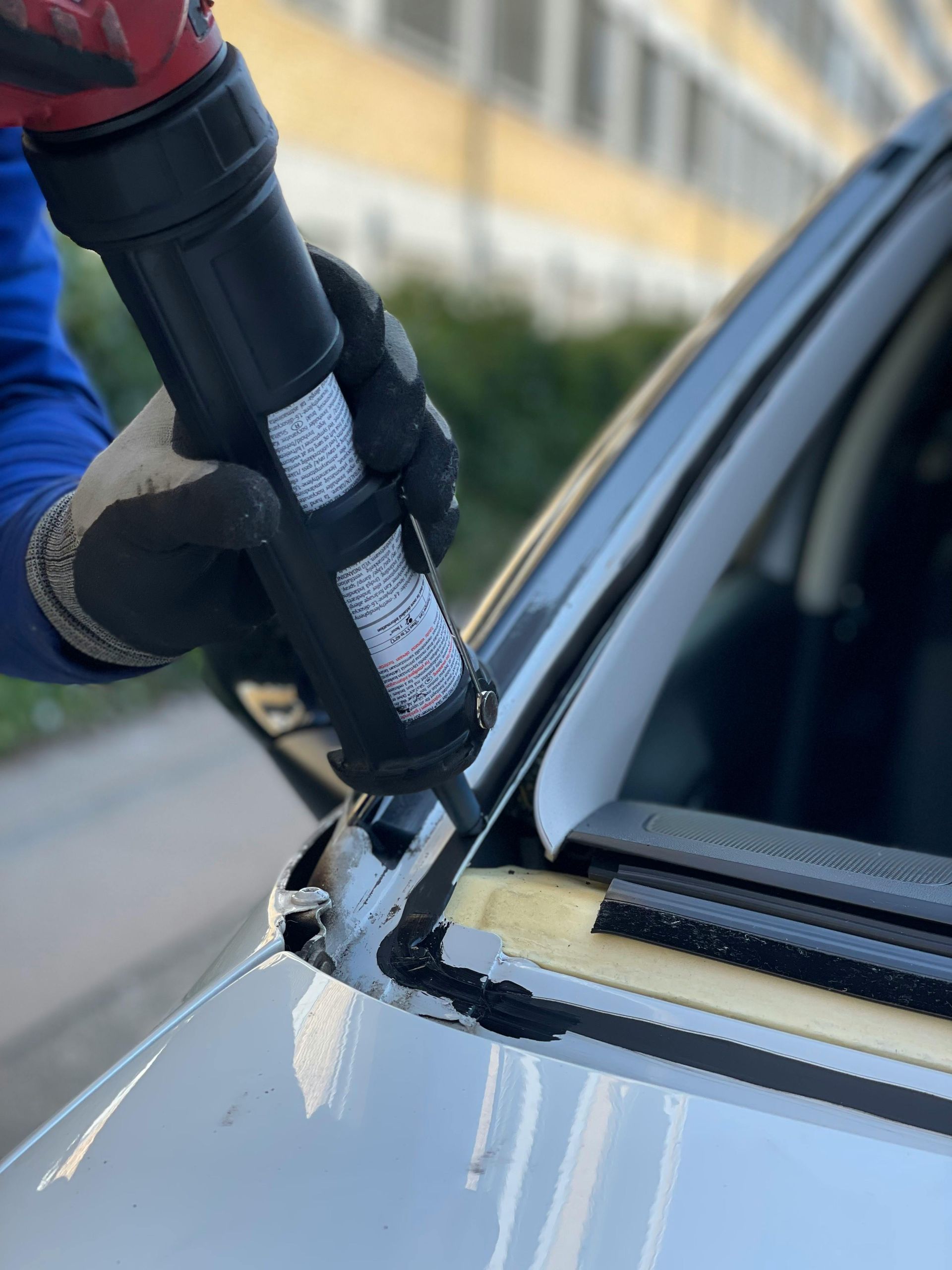 Person applying black sealant around a car's windshield with a caulking gun, outdoors.