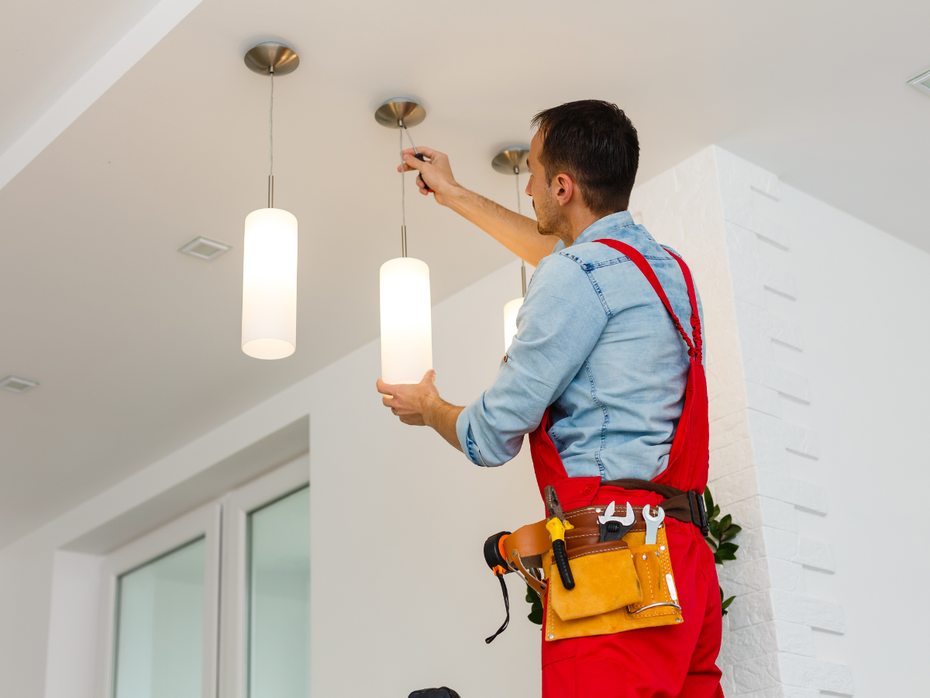 Electrician installing pendant lights on a white ceiling; wears red overalls and a tool belt.