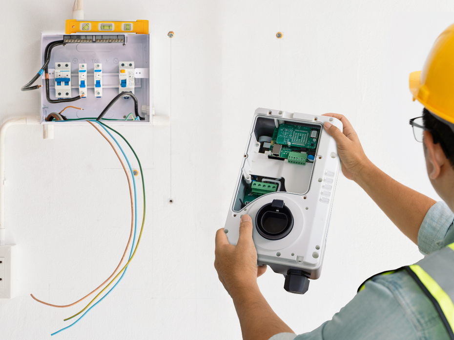 Electrician installing equipment on a wall, wiring visible, wearing a hard hat and safety vest.