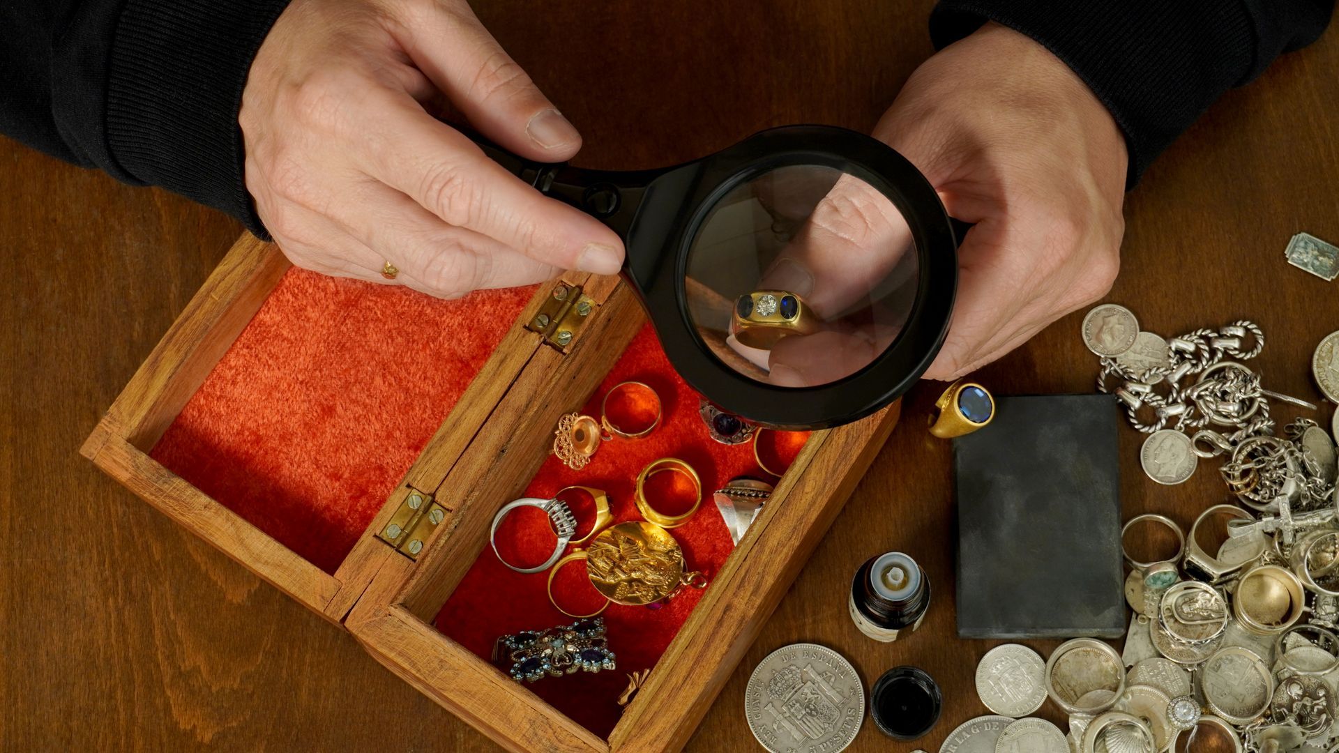 Hands holding a magnifying glass over an open wooden box lined various rings and jewelry.
