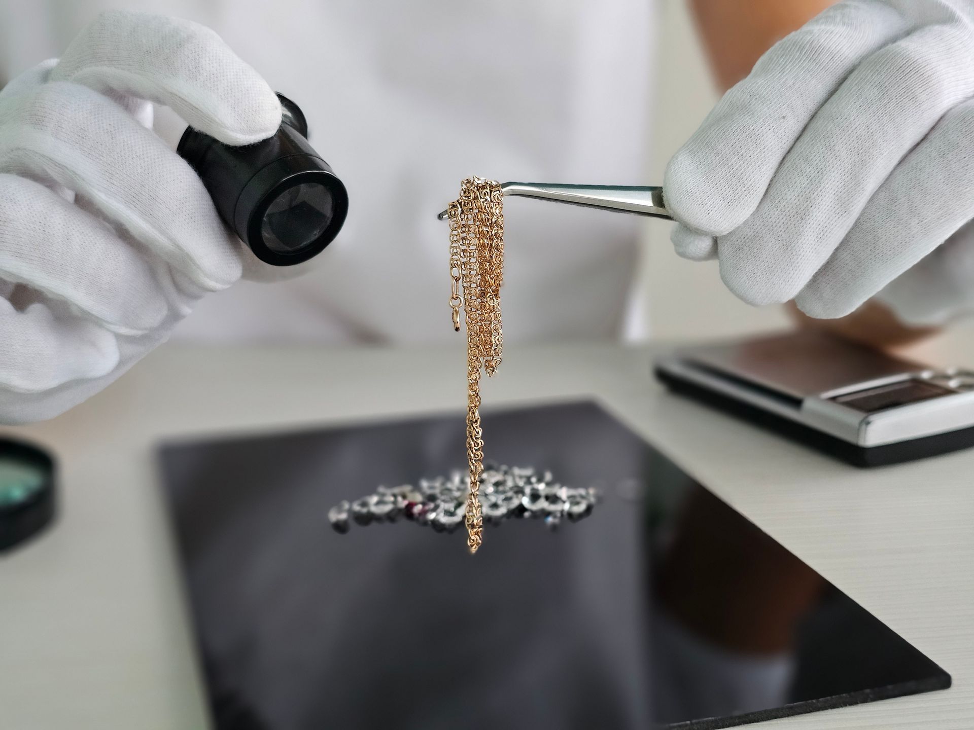 Appraiser inspects gold chain with magnifier and gloves beside jewelry and digital scale. Appraiser inspects gold chain with magnifier and gloves beside jewelry and digital scale.