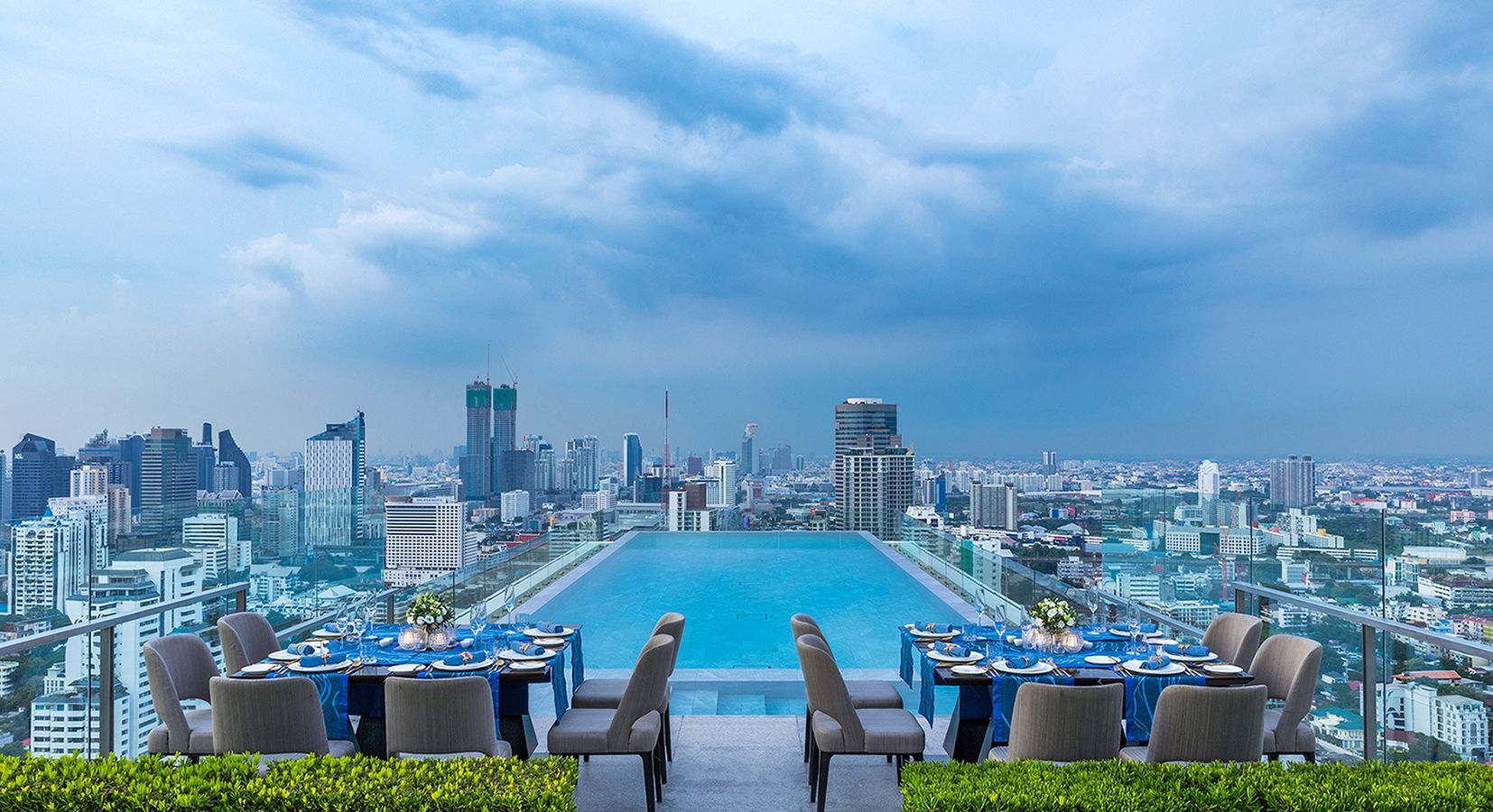 Rooftop dining area with tables set by a long swimming pool overlooking a city skyline under a cloudy sky.