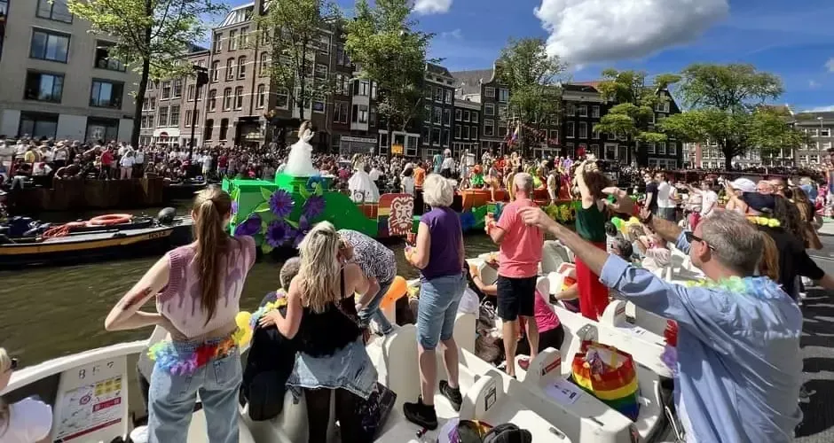 People watch a colorful boat parade along an Amsterdam canal on a sunny day.