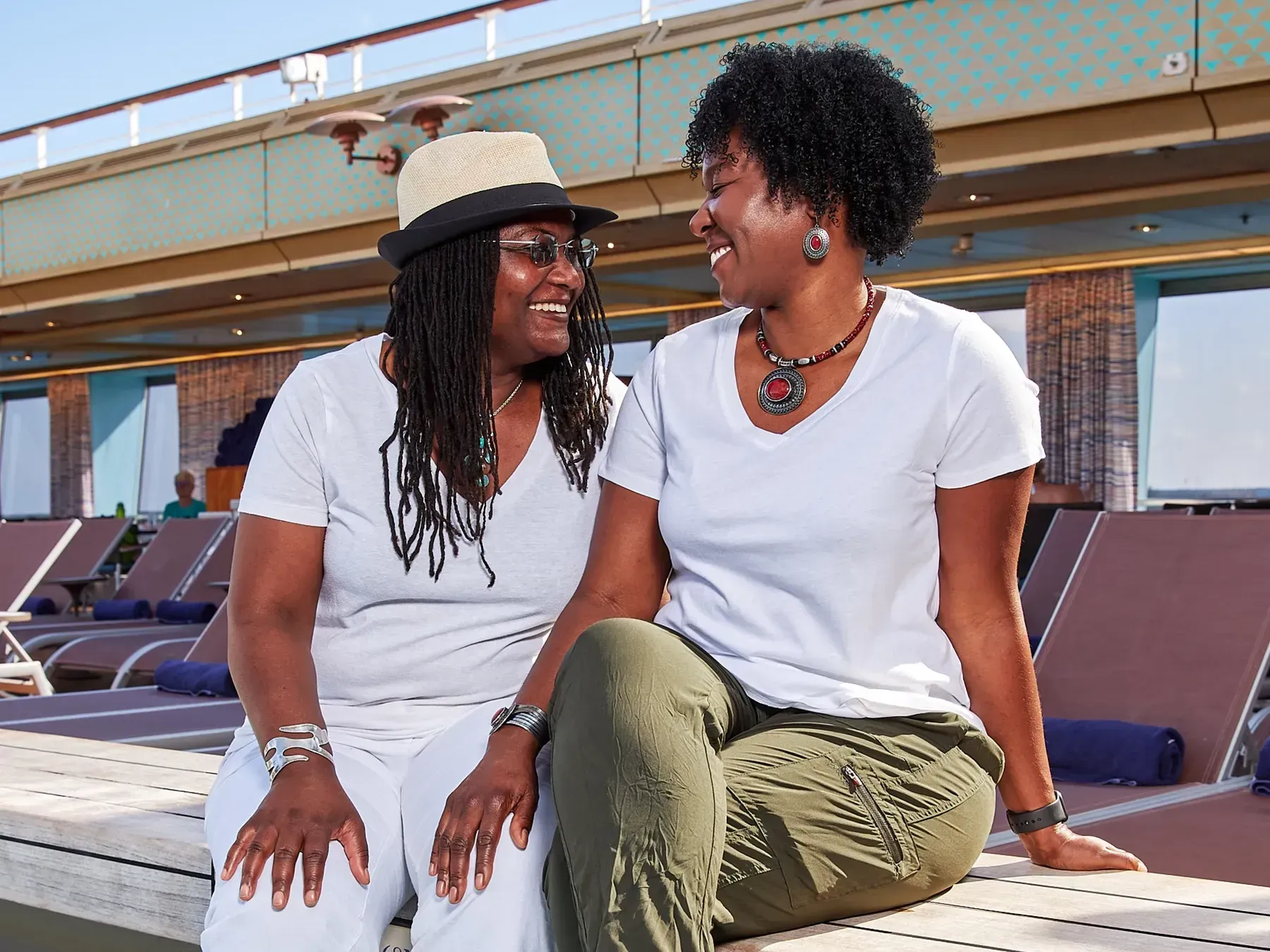 Two women smiling and looking at each other while sitting on lounge chairs on a cruise ship deck.