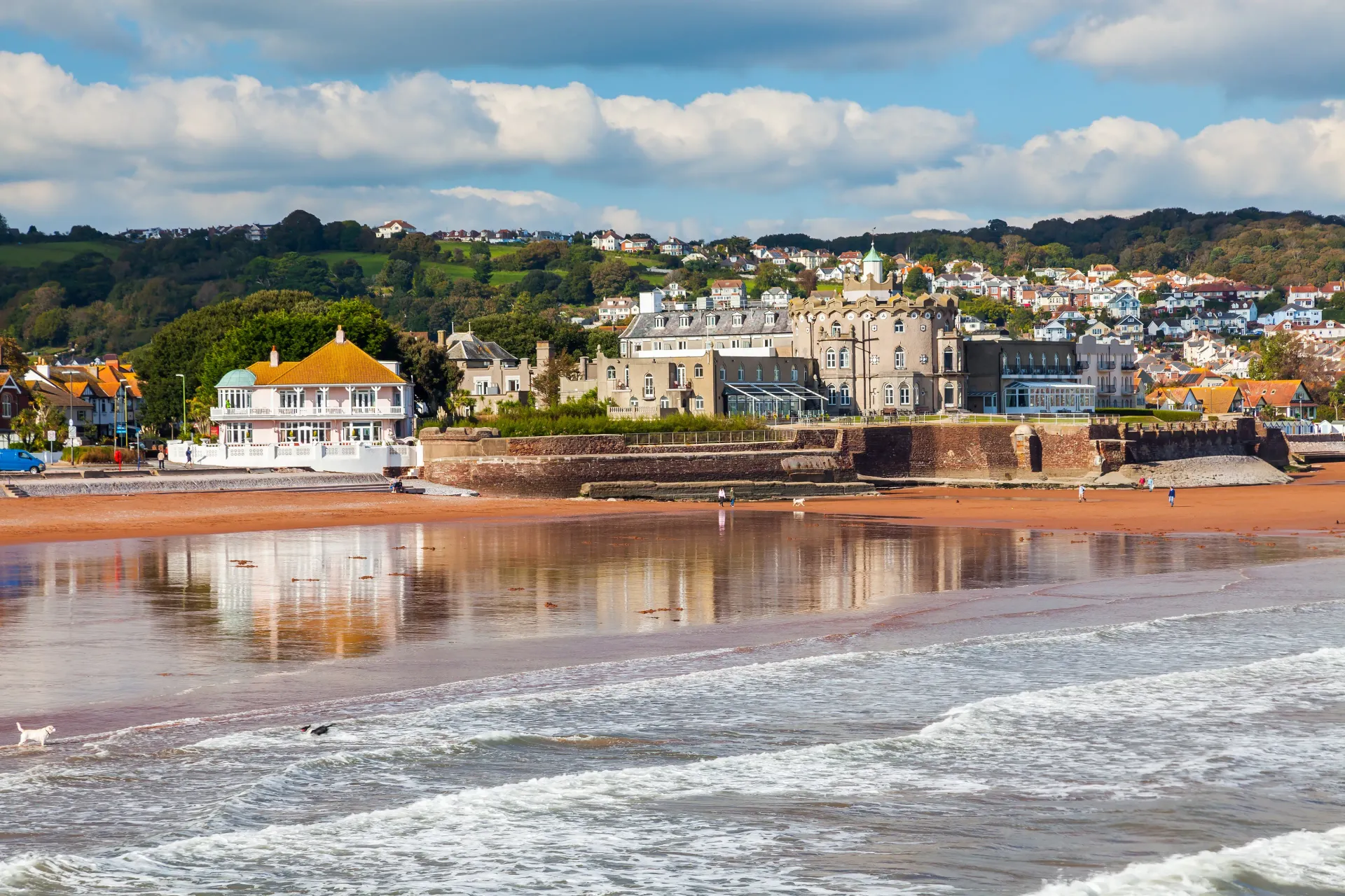 Coastal town with buildings, red sand beach, and blue sky.