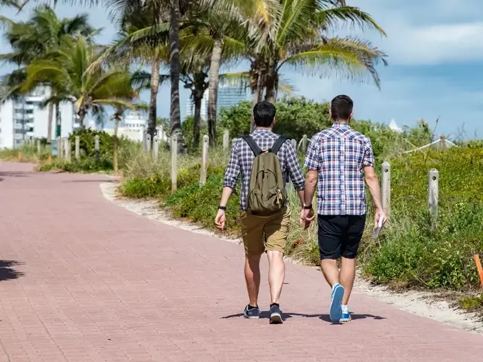 Two people walk hand-in-hand along a red path, palm trees in the background. Sunny day.