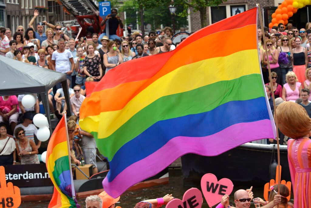 A large pride flag waves over a crowded Amsterdam canal during a pride parade, with people celebrating in the background.