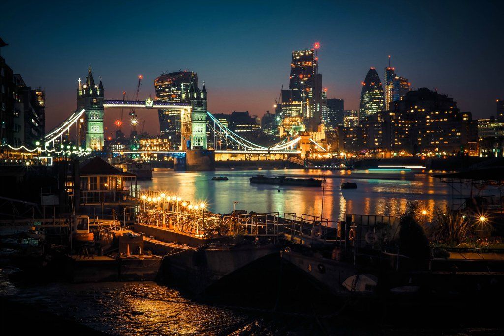 London skyline at night with Tower Bridge illuminated over the Thames River. City lights reflect on water.