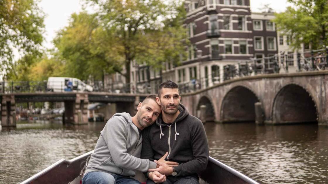 Two men in a boat on a canal in Amsterdam, one rests head on the other's shoulder, holding hands.