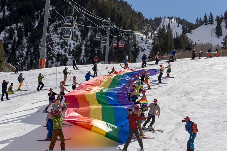 Skiers holding a rainbow flag on a snowy mountain slope; ski lift in the background.