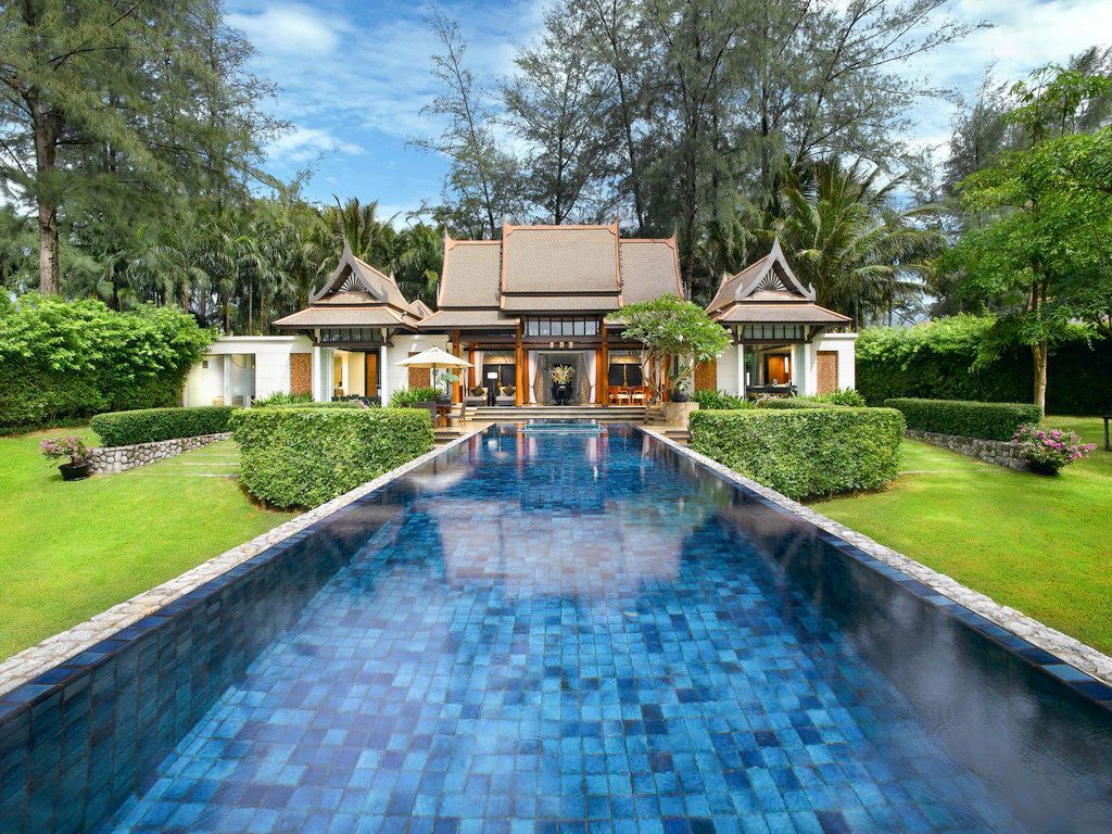 Luxury resort with a long blue pool leading to a traditional Thai-style building. Lush green lawn and trees surround.