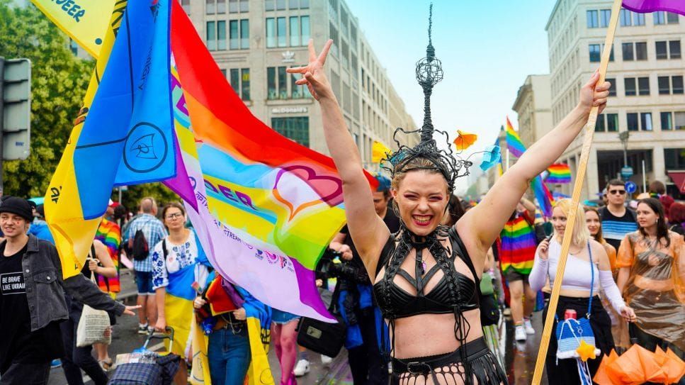 Woman with arms raised at a pride parade, holding rainbow flags in a city setting.