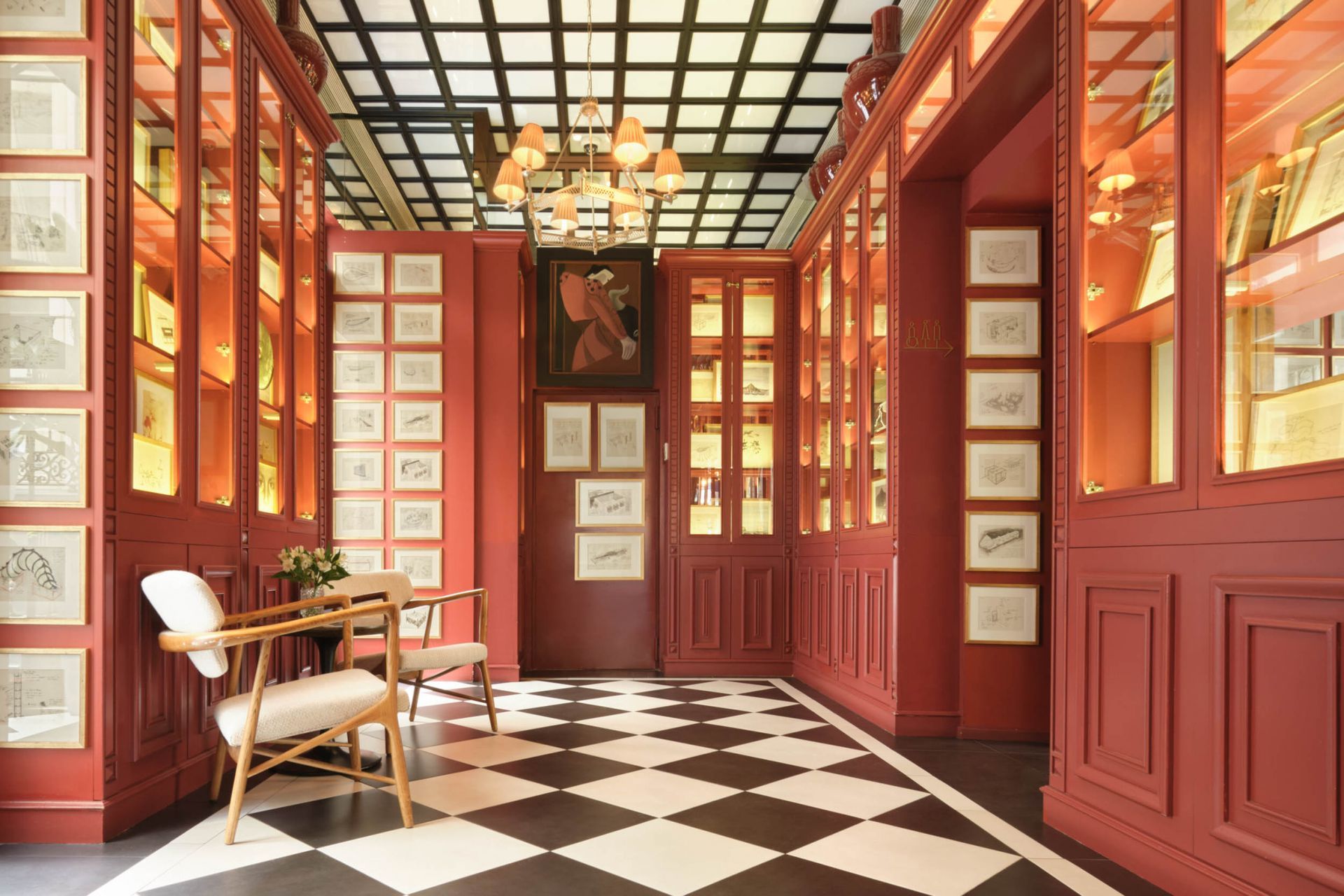 Red-walled library hallway with black and white checkered floor, glass display cases with artwork, two chairs.