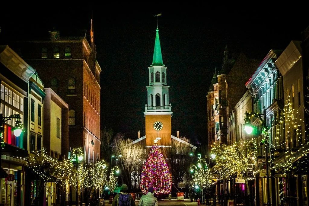 Night view of a street with a lit Christmas tree and church steeple, decorated with holiday lights. Burlington, Vermont
