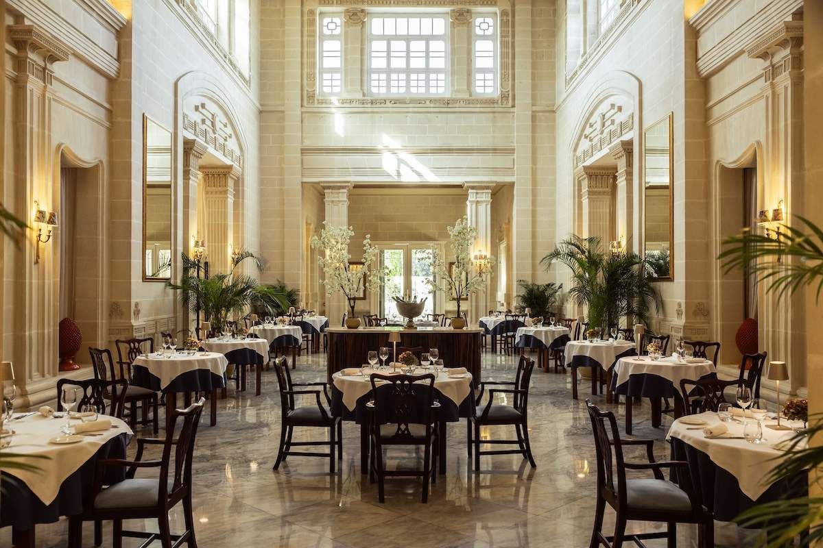 An elegant, sunlit dining hall with marble floors, classical columns, and numerous tables set for fine dining.