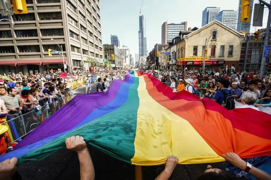 Rainbow flag held by people in a street parade, Toronto, Ontario. Buildings and crowd in the background.