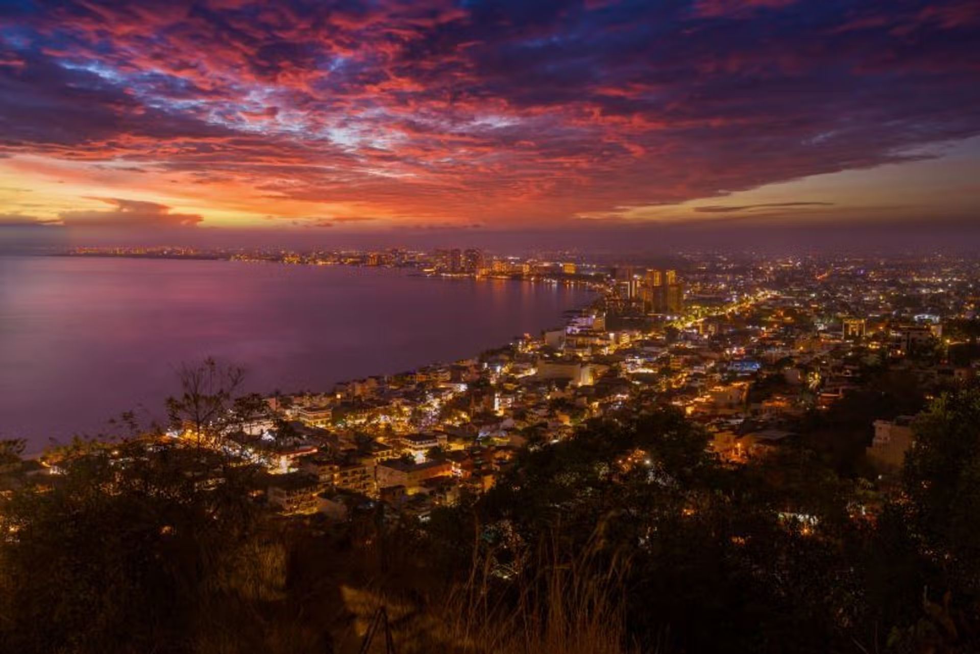 City skyline at dusk with lights, set along a bay. Dramatic sunset colors of pink, purple, and orange in the sky.