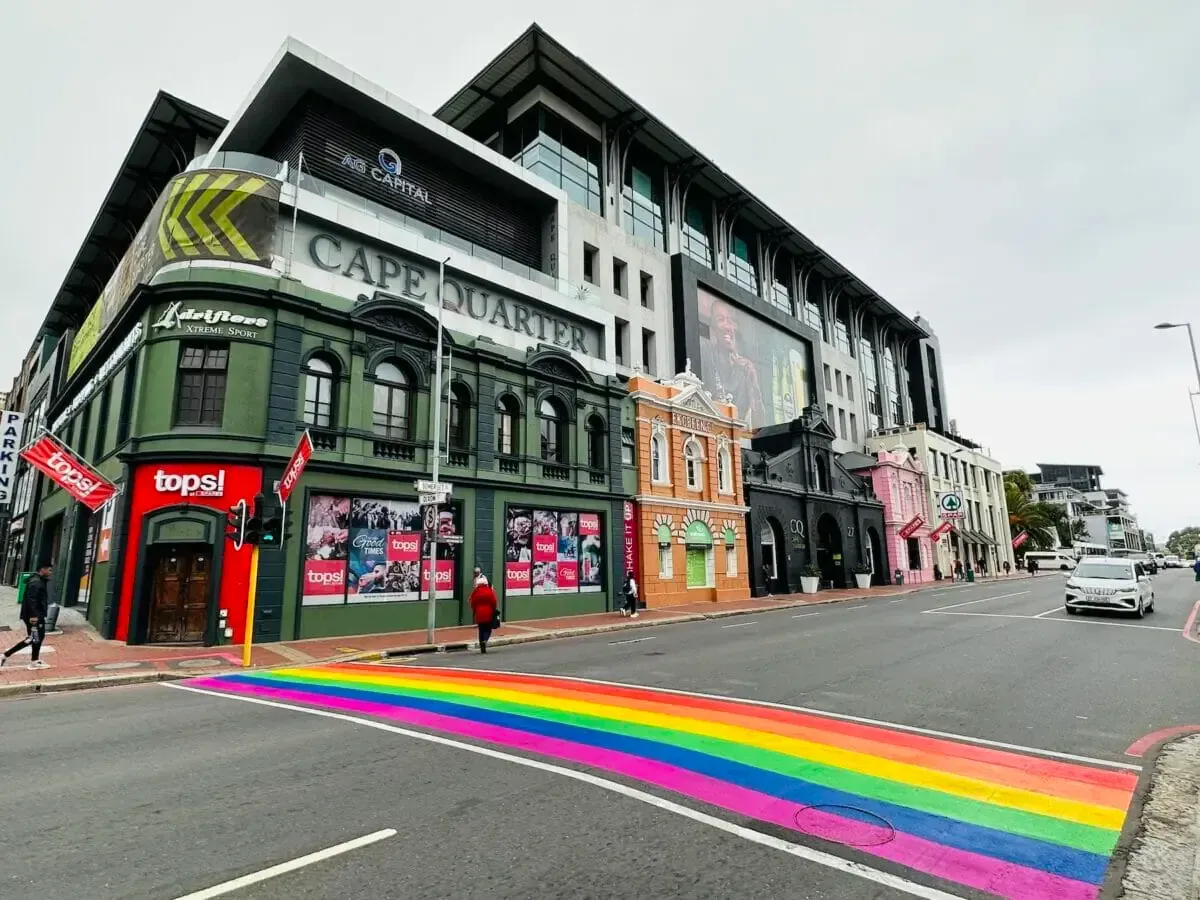 Street with rainbow crosswalk in front of colorful buildings, including a Cafe Quarter. A few people and a car are present.