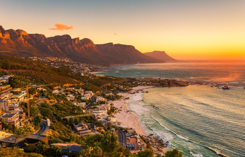 Coastal view of Cape Town, South Africa, at sunset. Mountains, beach, and ocean are bathed in orange and gold light.