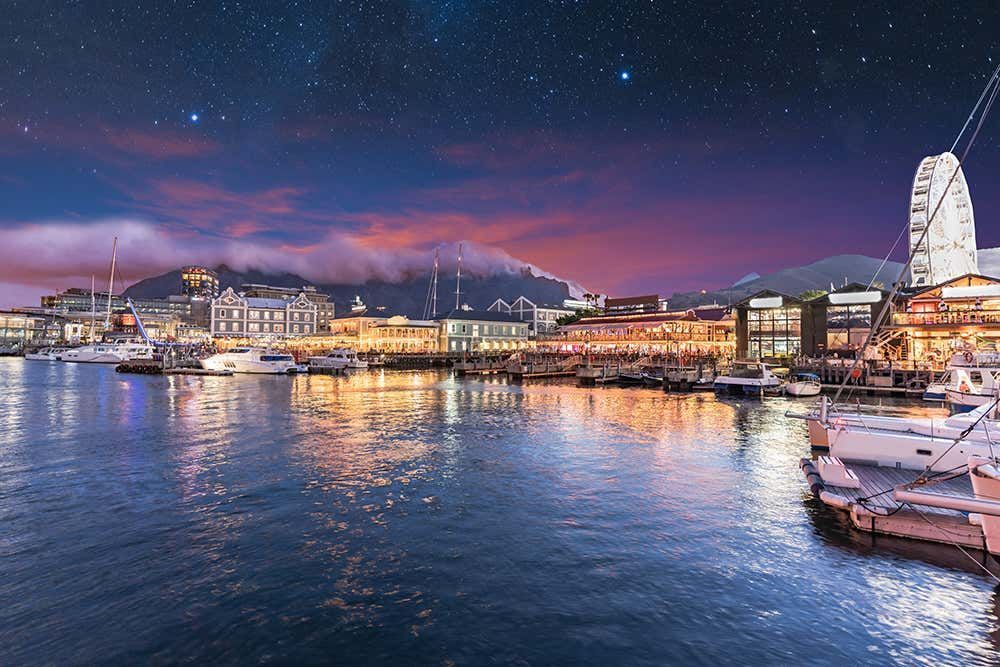 Waterfront at dusk with Ferris wheel, boats, and buildings under a starry sky. Cape Town
