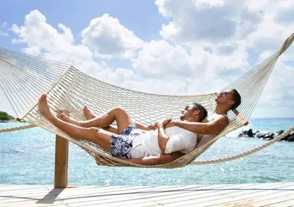 Two people relax in a hammock by the ocean. Blue sky, water, and white hammock.