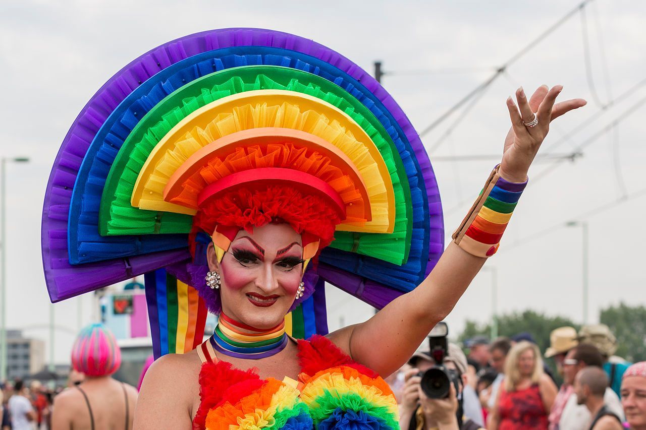 Drag queen in rainbow attire at a Pride parade, arm raised, smiling.