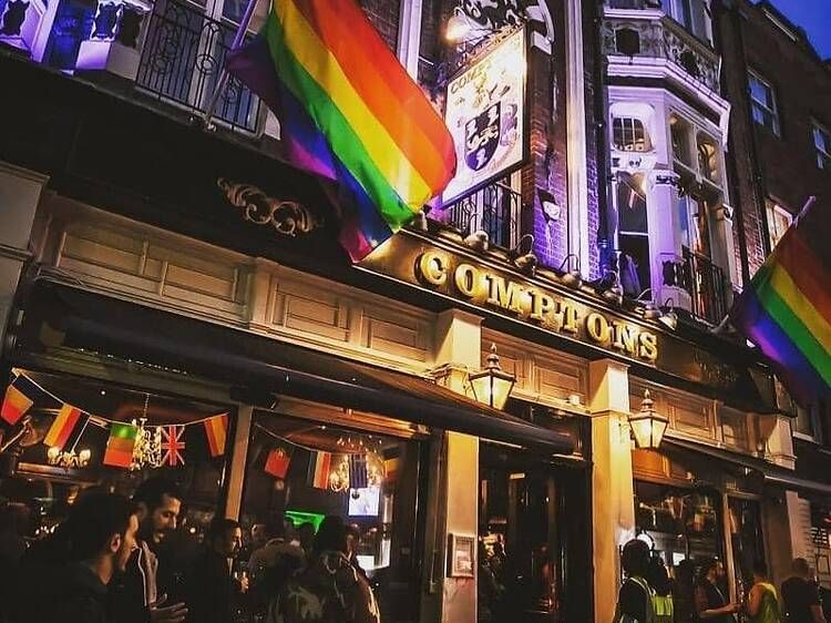 Exterior of Compton's, a pub with rainbow flags. People gather outside, illuminated by lights.