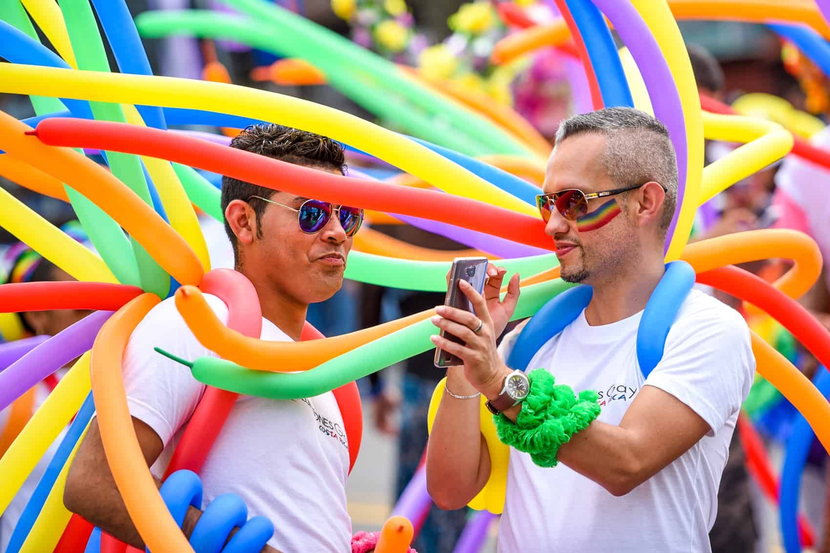 Two people in white shirts and sunglasses smiling amid colorful balloon tubes at an outdoor event