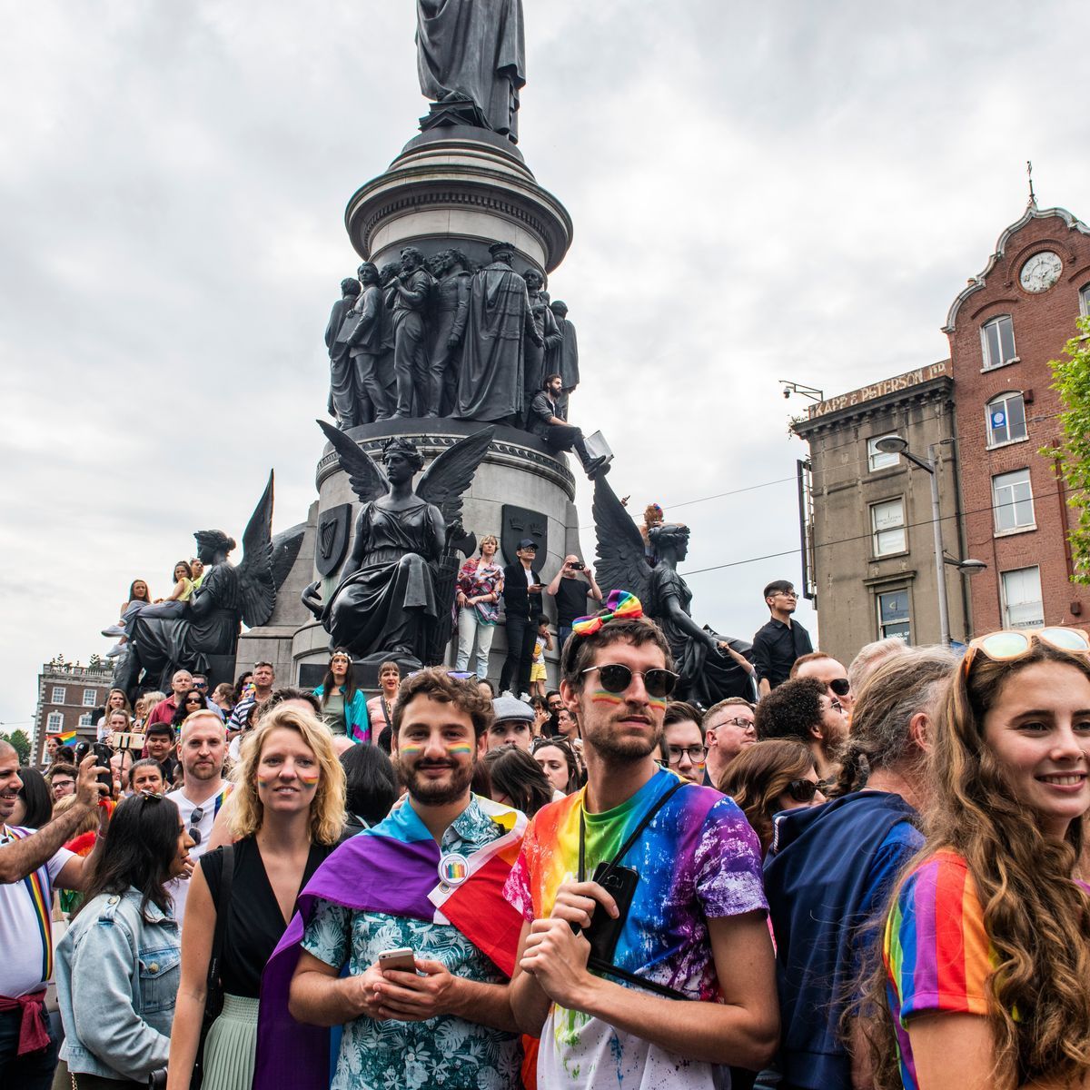 A crowd celebrates at a pride event in front of a tall, ornate stone monument in a city plaza.