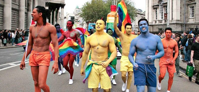 A group of people wearing colorful body paint and matching shorts march in a parade while holding rainbow flags.