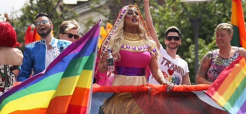 People celebrate at a Pride parade, waving rainbow flags while some wear vibrant, expressive outfits and accessories.