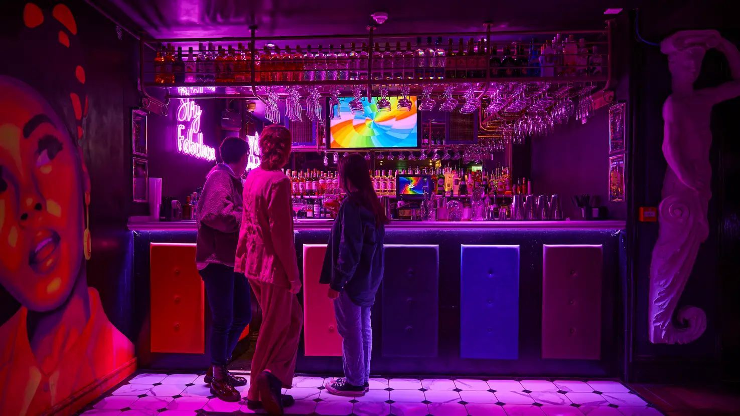 Three people stand at a neon-lit bar with colorful panels, under a glass rack, in a room with a large mural.