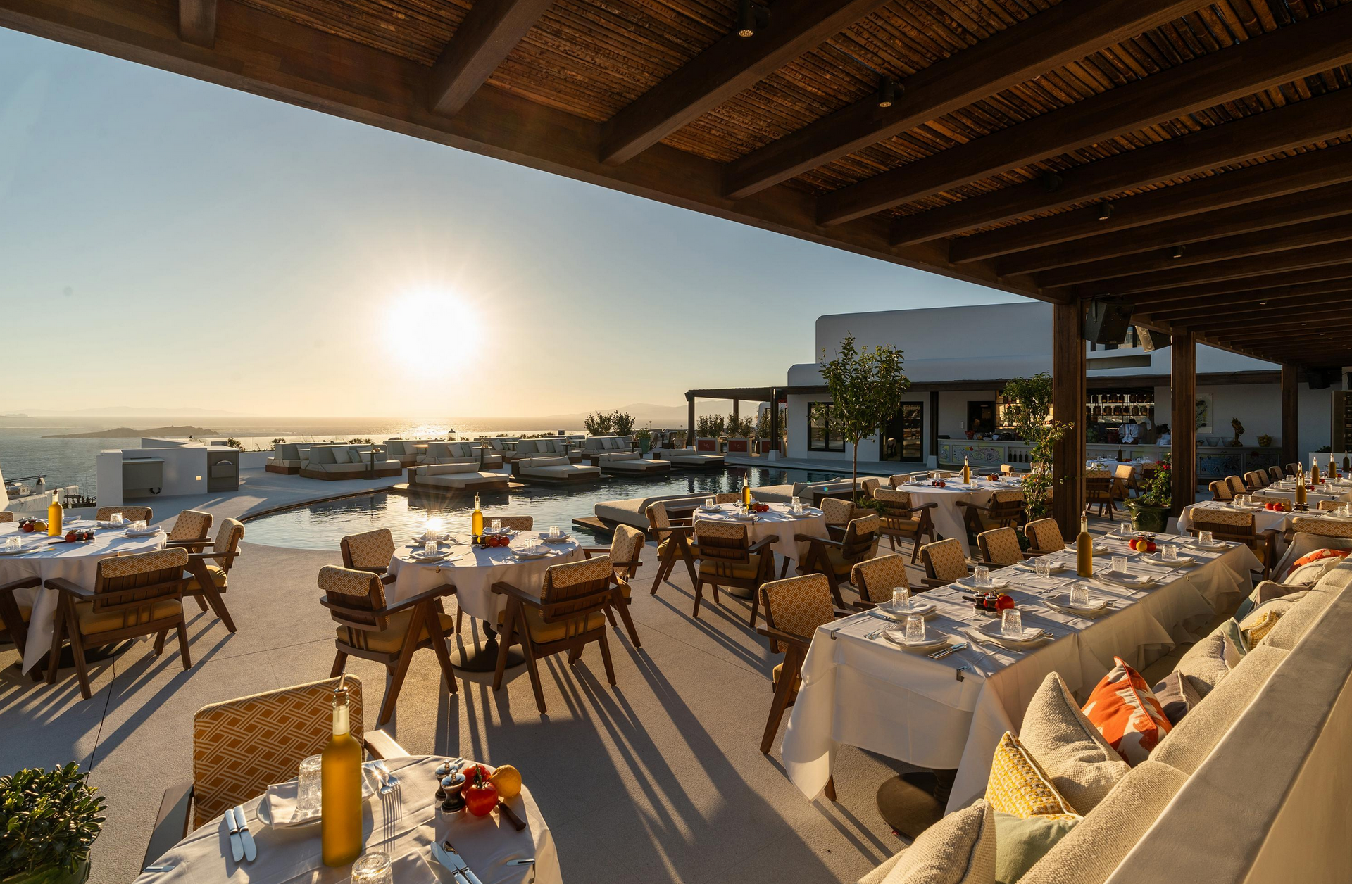 Outdoor restaurant with tables set for dinner, overlooking a pool and ocean at sunset. Elysium Hotel Mykonos