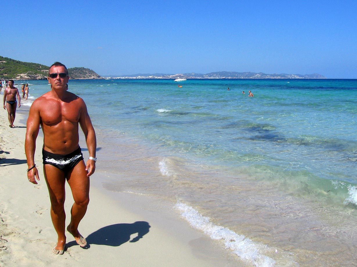 Man with muscular physique walking on a sandy beach. Clear turquoise water, sunny day. Es Cavallet, Ibiza