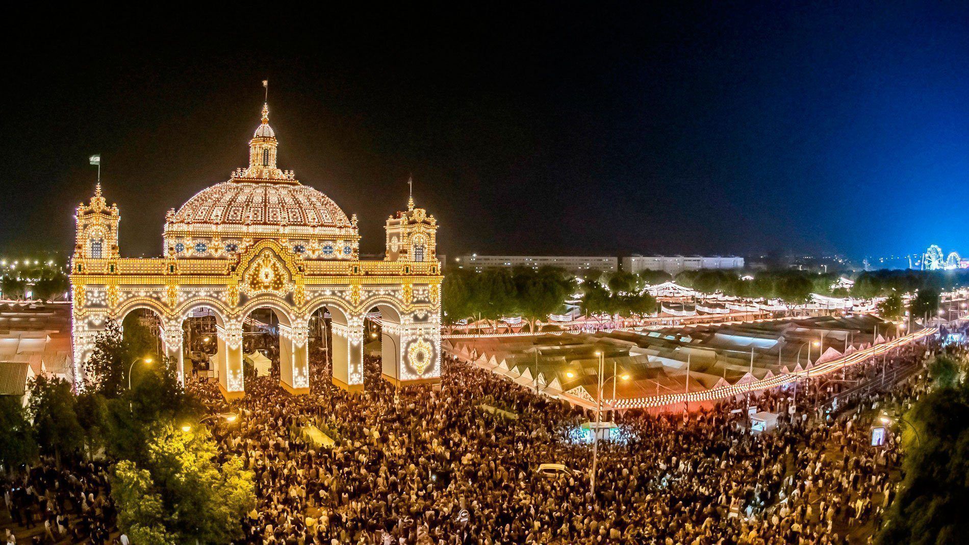 Night view of a brightly lit fair archway with crowds, lights, and buildings in the background. Feria de San Miguel in Seville Spain