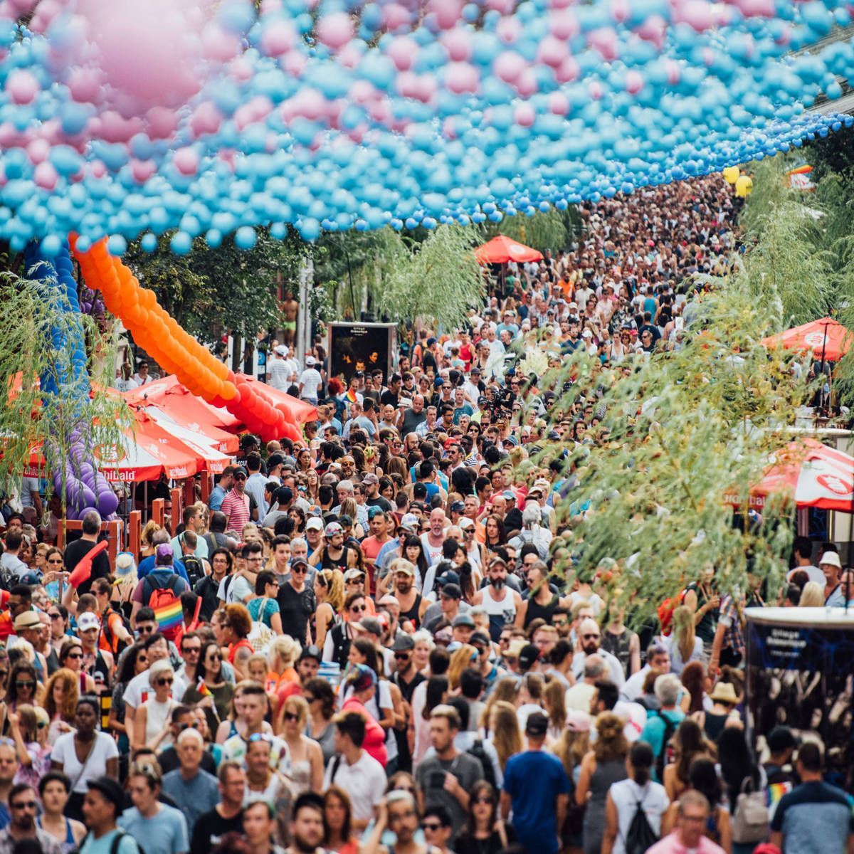 A large crowd of people walking on a tree-lined street under blue and pink decorations; red umbrellas are visible. Fierté Montréal