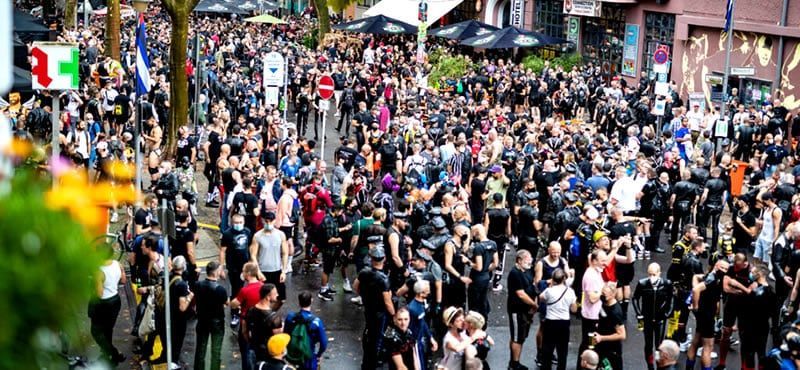 A large crowd of people on a city street, many wearing black. Some are gathered near outdoor cafes. Folsom Berlin