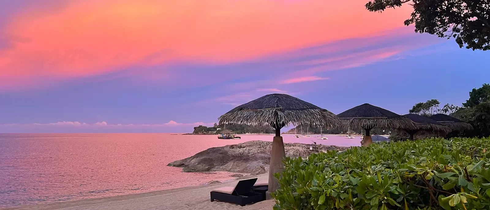 Sunset over a beach with pink and purple sky, lush green foliage, and two dark lounge chairs.