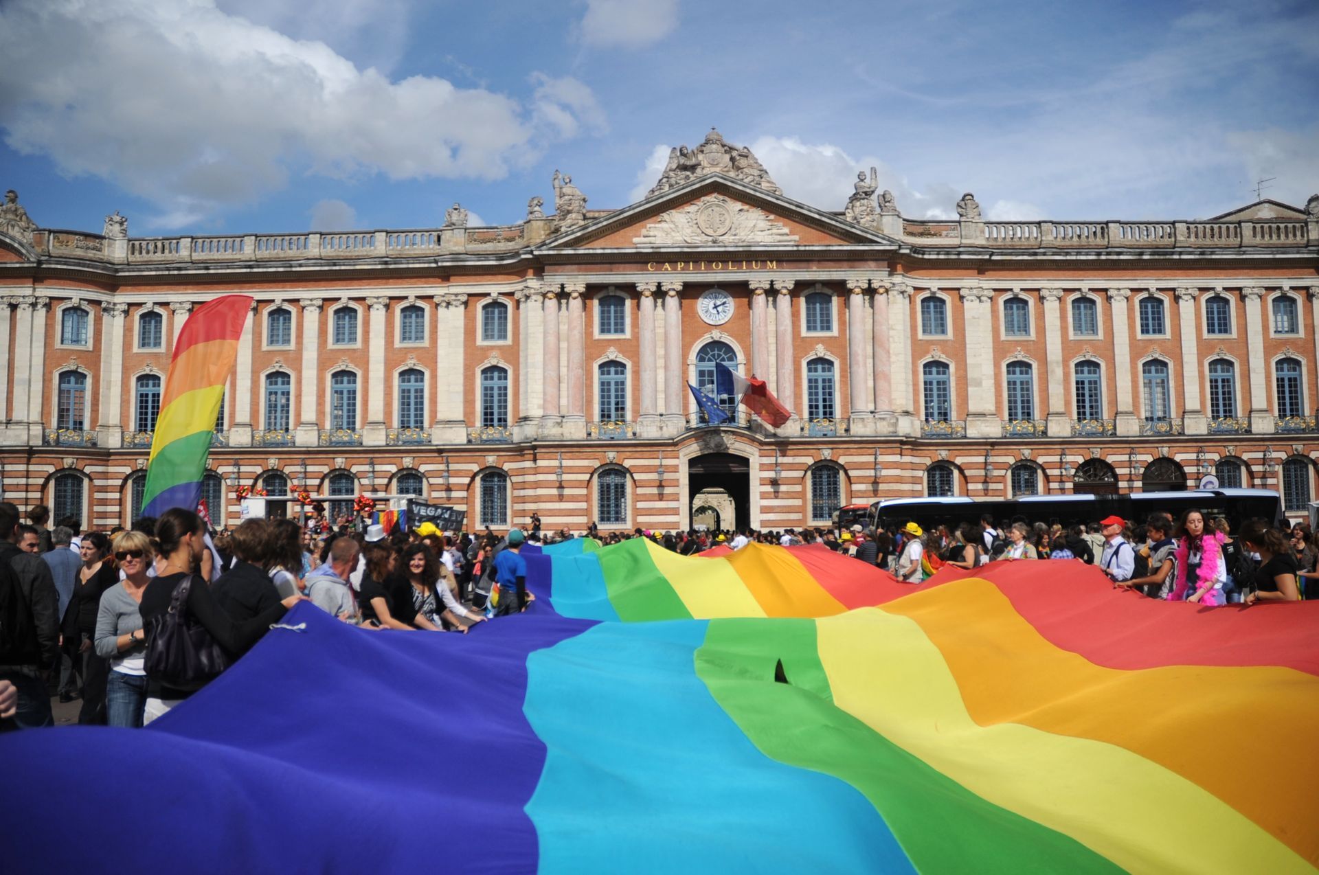 Large rainbow flag held by people in front of a building with a Pride flag, sunny day.