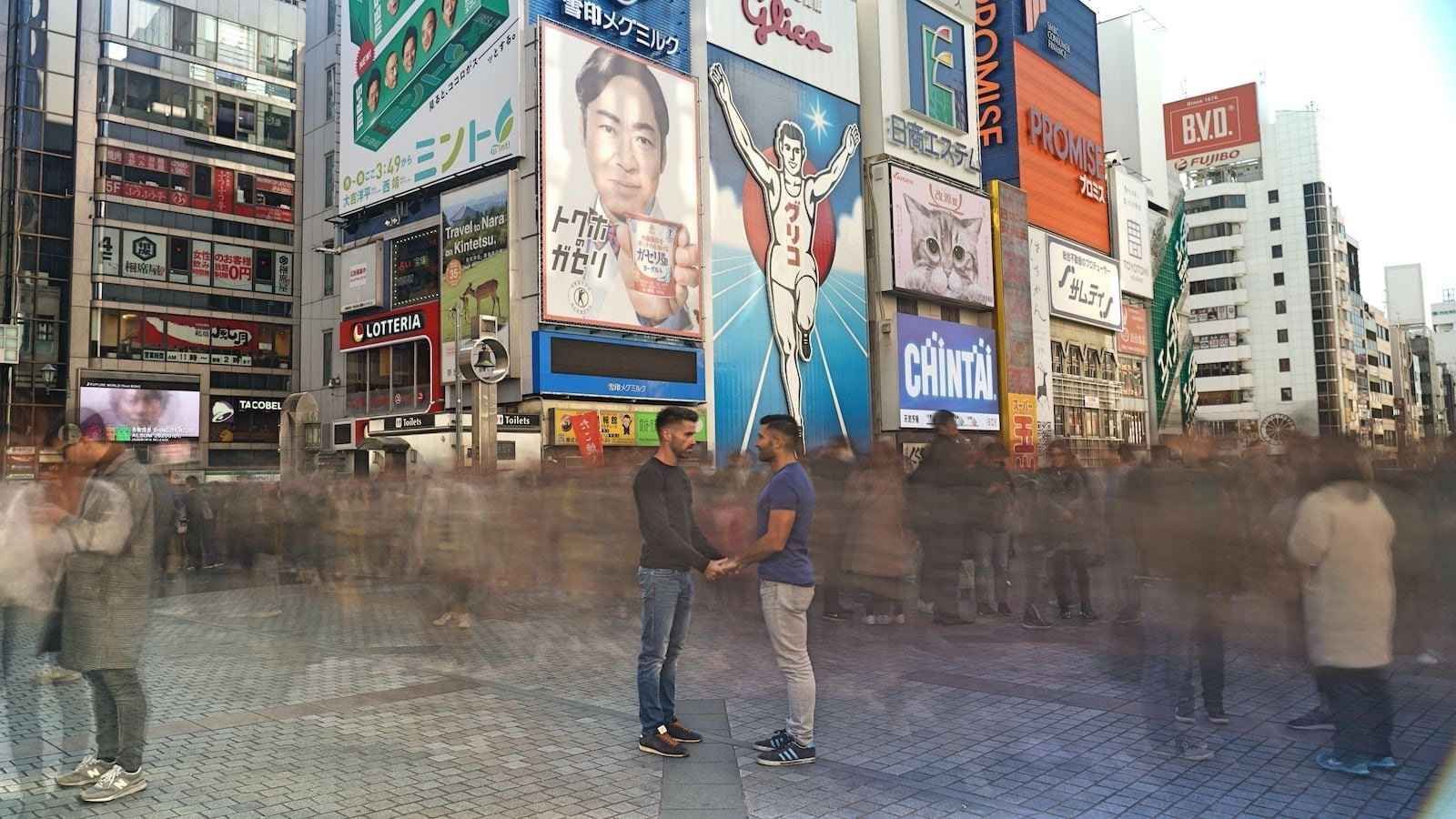 Two men holding hands in a crowded city square with illuminated billboards.