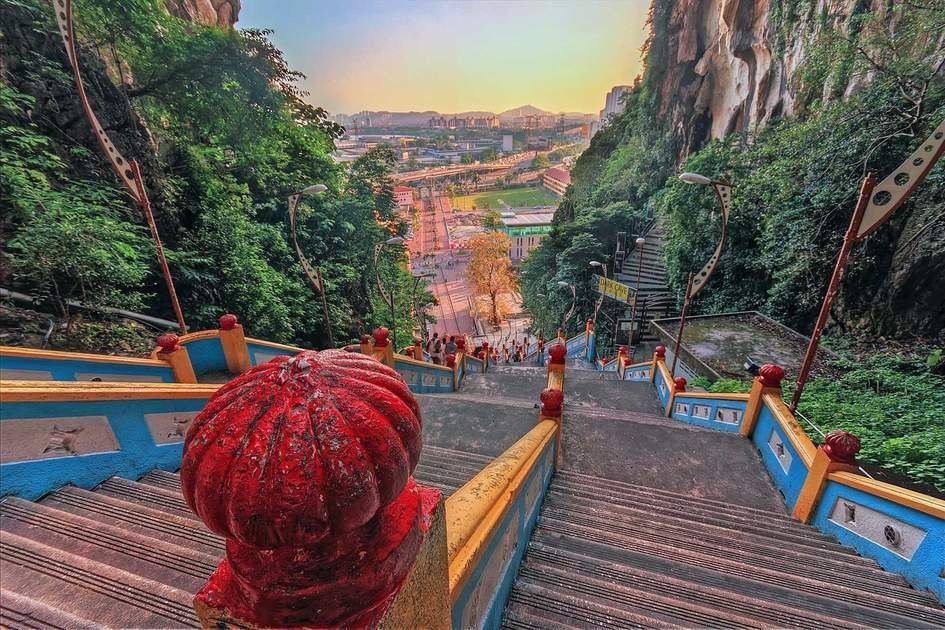 Stone steps leading down from a temple with red railings, lush greenery on either side, and a city in the background.