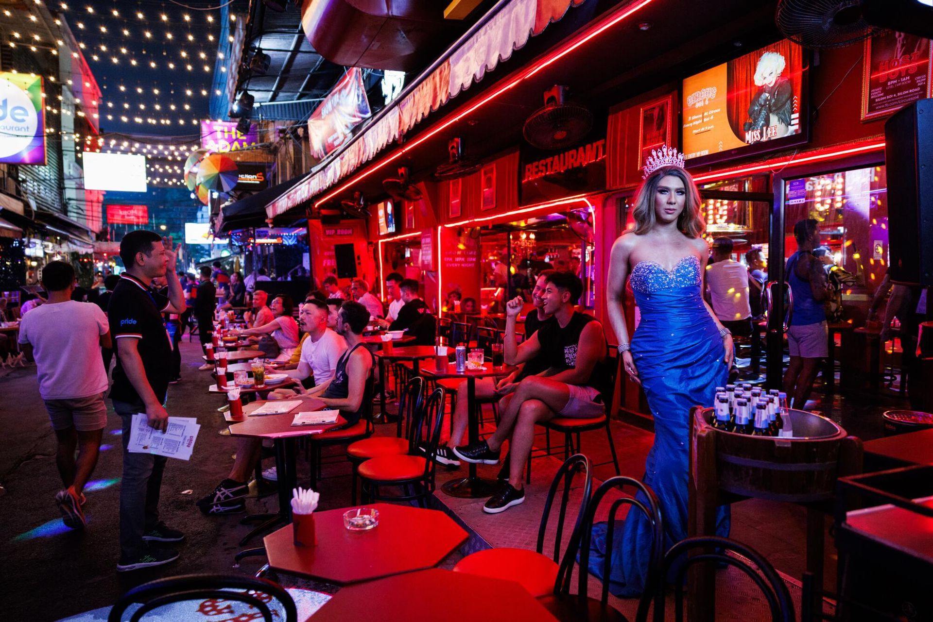 Street scene with people seated at tables outside bars, bright red and neon lights. A person in a blue dress stands on a stand.