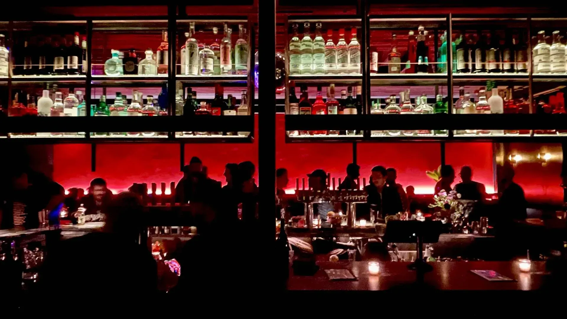 Bar scene, dimly lit with red accent lights, silhouettes of people at tables and bar. Shelves of bottles. Gay Bar