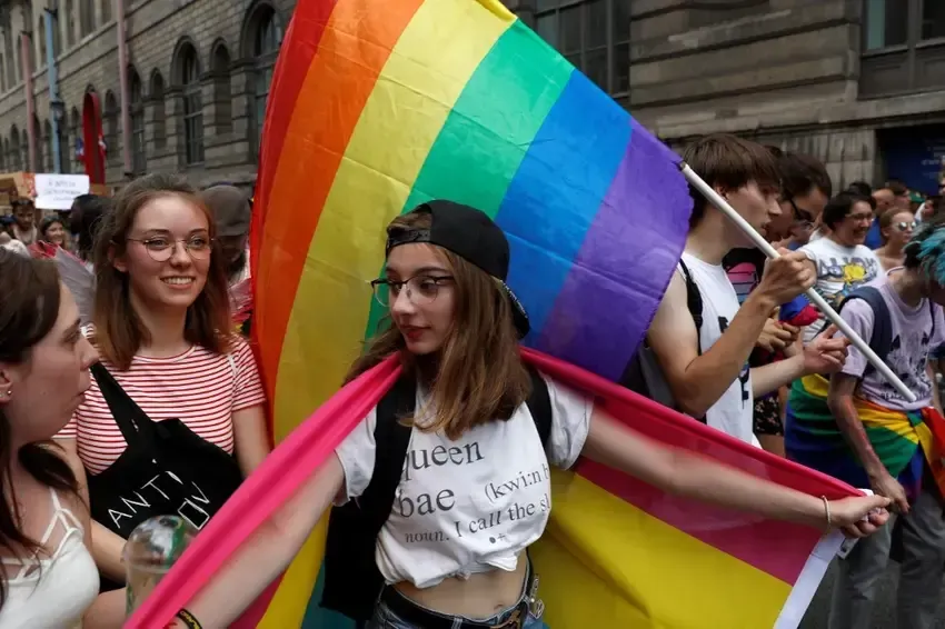 People at a Pride parade; one with rainbow flag draped over, wearing a black cap. Others in crowd, holding flags.