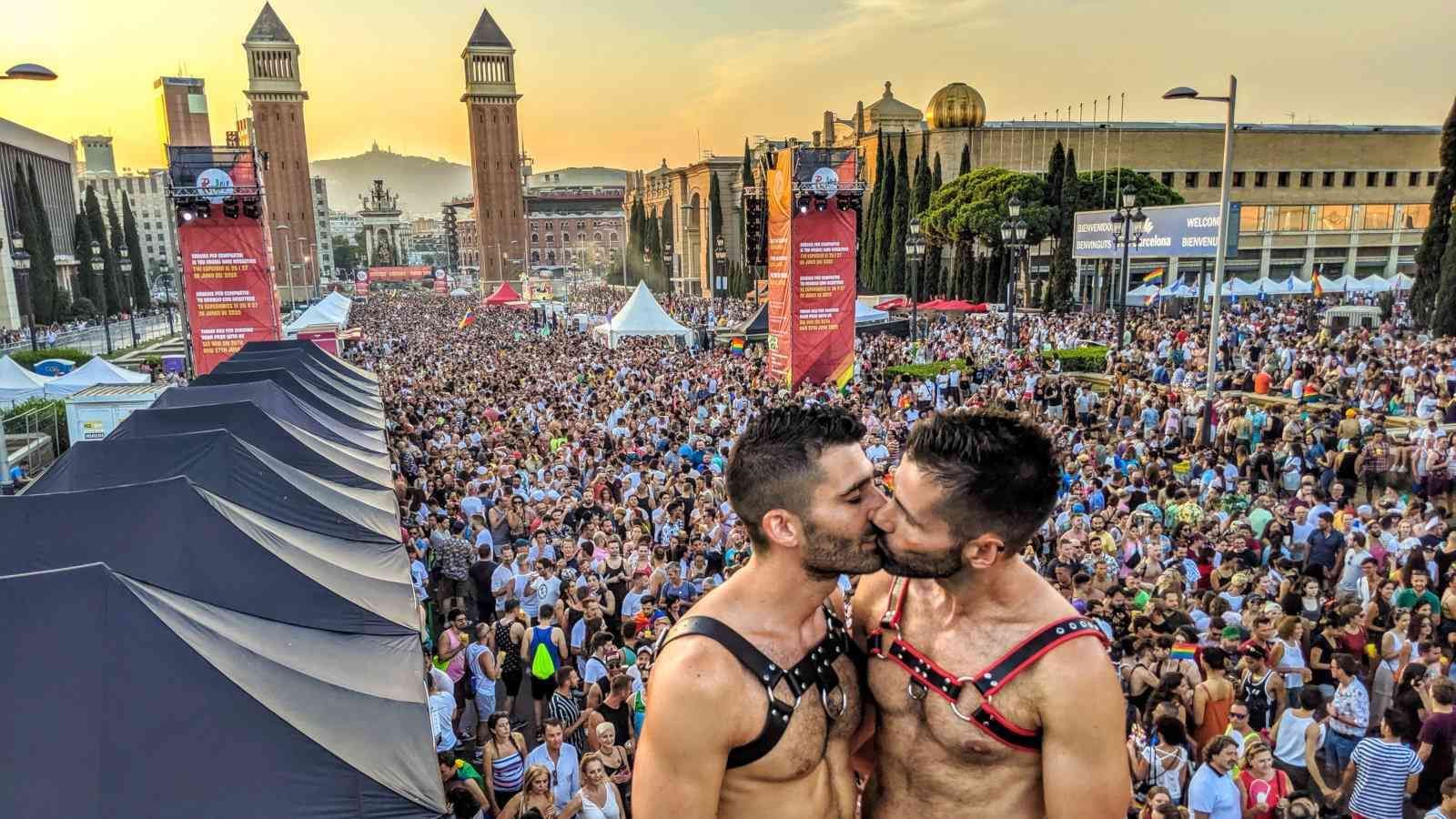 Two men kissing at a crowded outdoor event. Sunset in the background; buildings and banners visible. Barcelona pride
