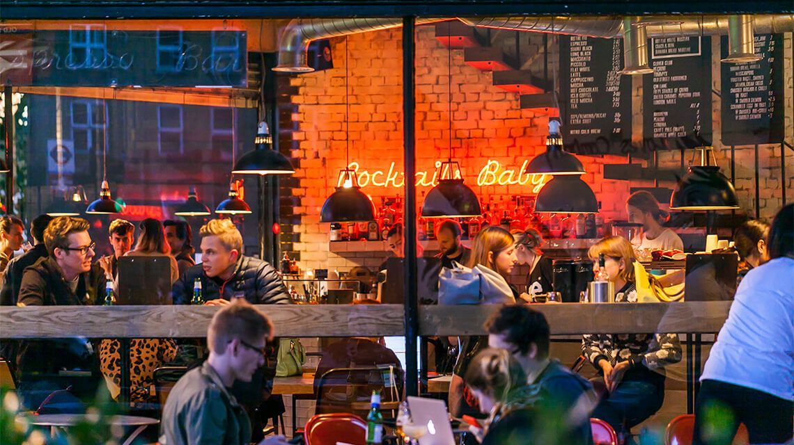 Cafe interior with patrons at tables using laptops, illuminated by neon lights and hanging lamps.
