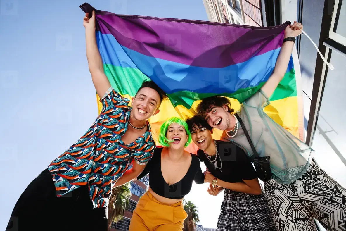 Four smiling people holding up a rainbow flag outdoors.